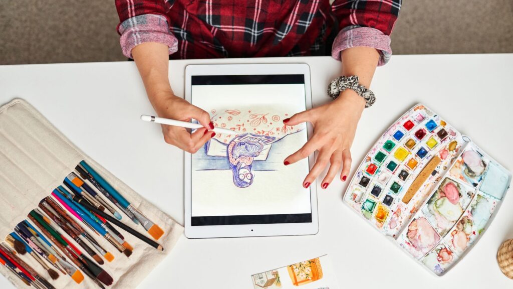 Artist drawing a colorful digital illustration on a tablet using a stylus, surrounded by watercolor paints and brushes on a white desk